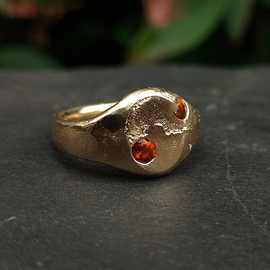 The 9ct Sandcast Sapphire Signet by silverhollowjewellery is a gold ring with a rough, organic texture and two round yellow and orange sapphires, shown on dark stone with a blurred green background.