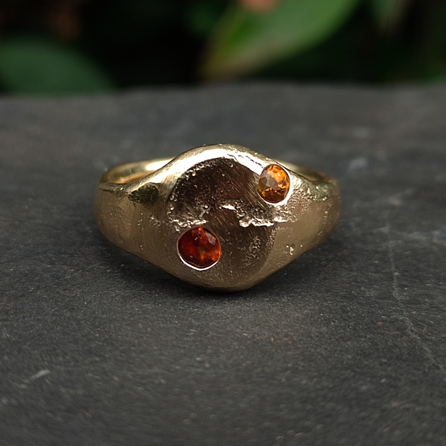 The 9ct Sandcast Sapphire Signet by silverhollowjewellery is handcrafted from recycled gold, featuring a textured surface and two asymmetrically set round yellow and orange sapphires, shown on a dark stone background.