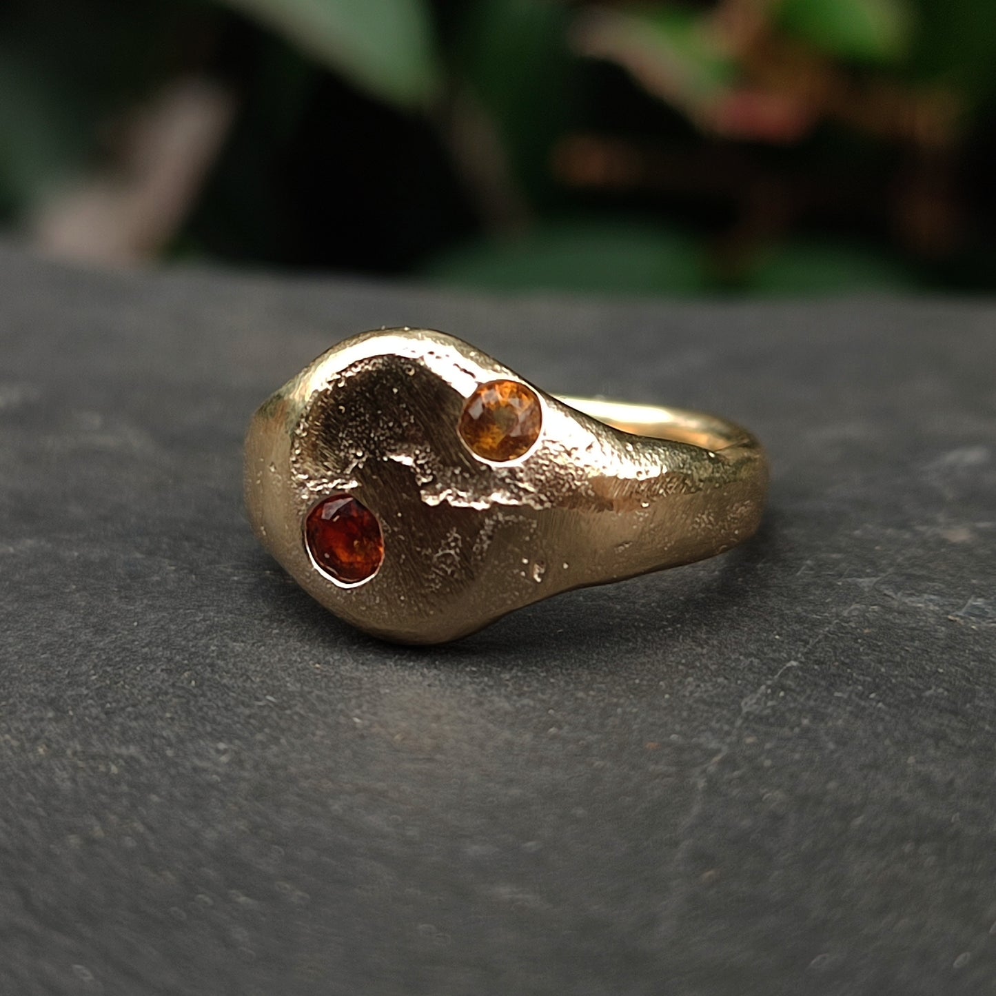 The 9ct Sandcast Sapphire Signet by silverhollowjewellery is a handcrafted gold ring with an organic shape, made from recycled gold and set with two small round yellow and orange sapphires in an asymmetric design.