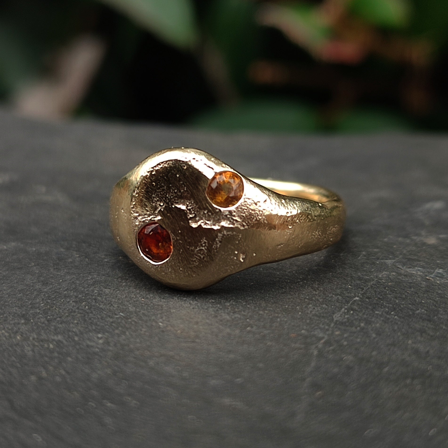 The 9ct Sandcast Sapphire Signet by silverhollowjewellery is a handcrafted gold ring with an organic shape, made from recycled gold and set with two small round yellow and orange sapphires in an asymmetric design.