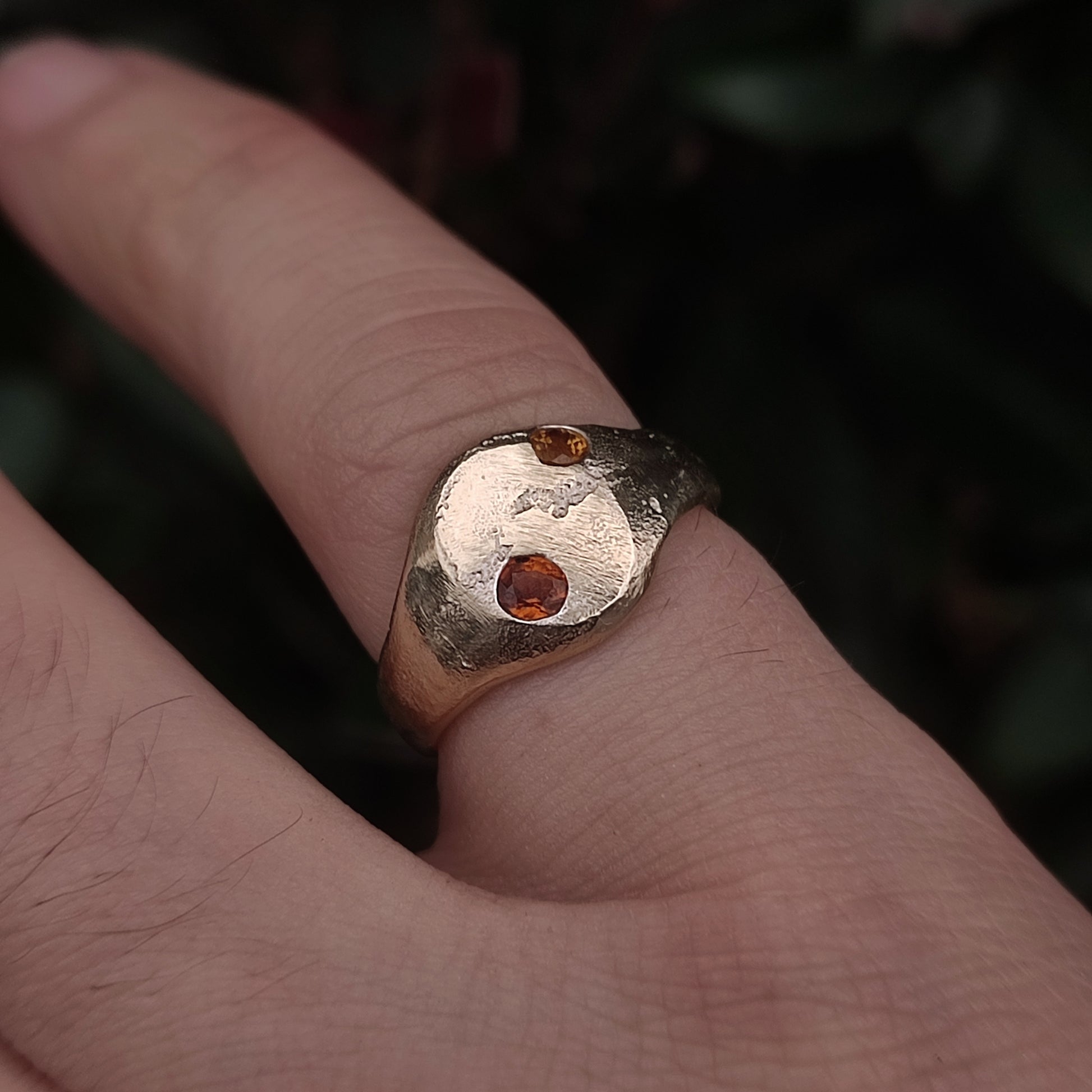 The 9ct Sandcast Sapphire Signet by silverhollowjewellery, featuring two small yellow and orange sapphires, is worn on a finger against a blurred dark background.