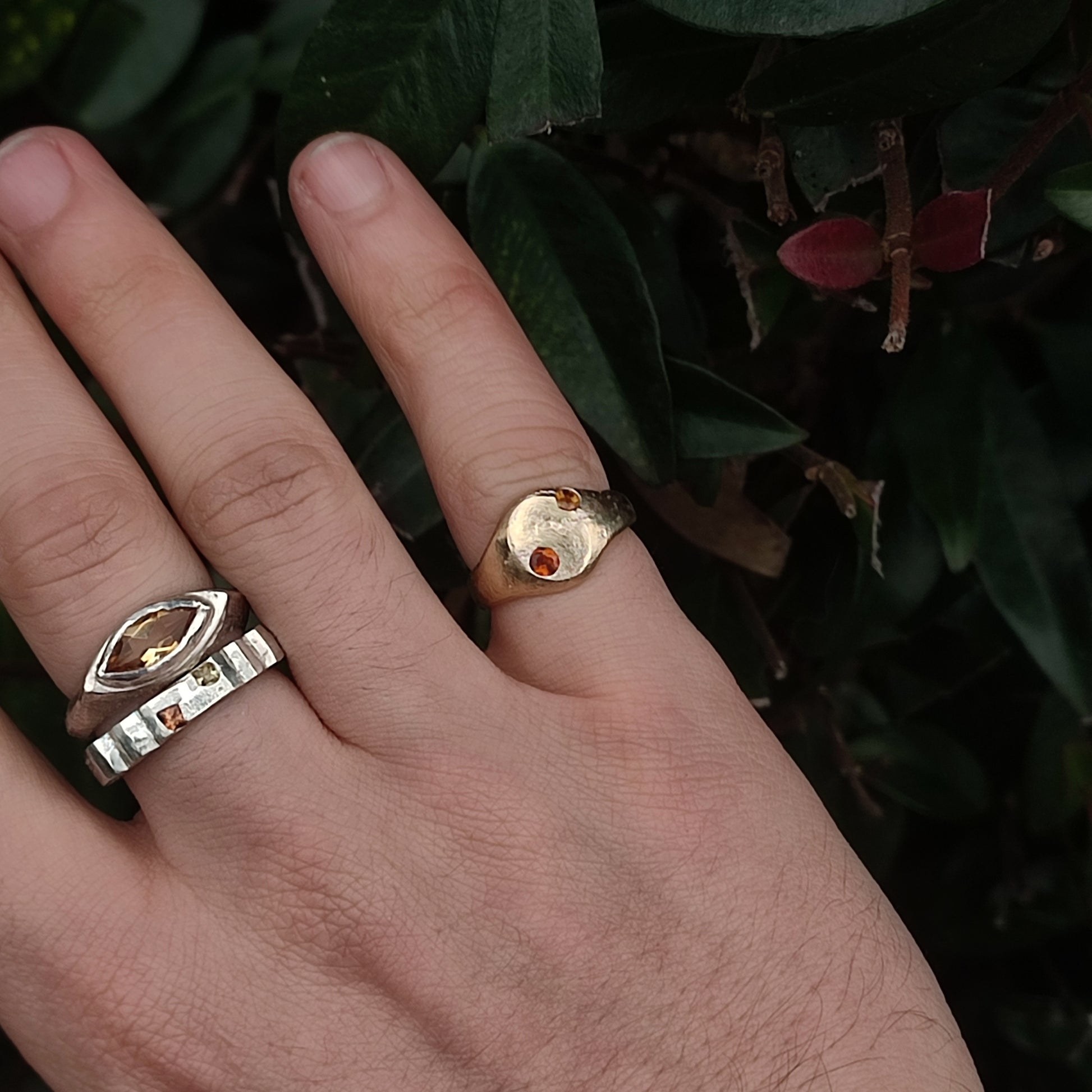 A hand wearing two silverhollowjewellery 9ct Sandcast Sapphire Signet rings, each set with yellow and orange sapphires, is held against green leaves.