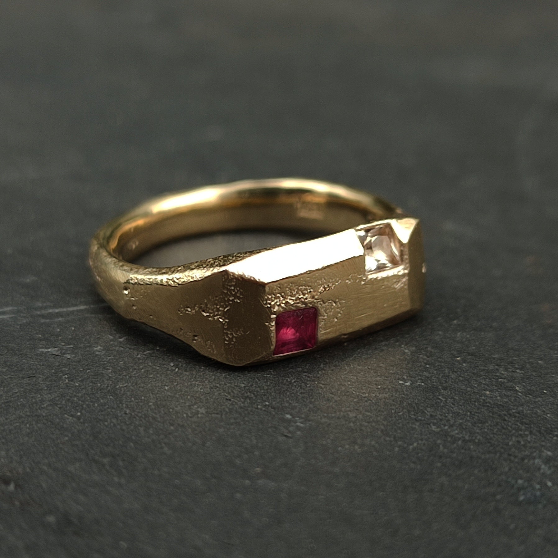 The 9ct Sandcast Sapphire/Ruby Signet by silverhollowjewellery is crafted from recycled gold using sand casting, featuring a textured band and square ruby and white sapphire set side by side on a rectangular bezel.