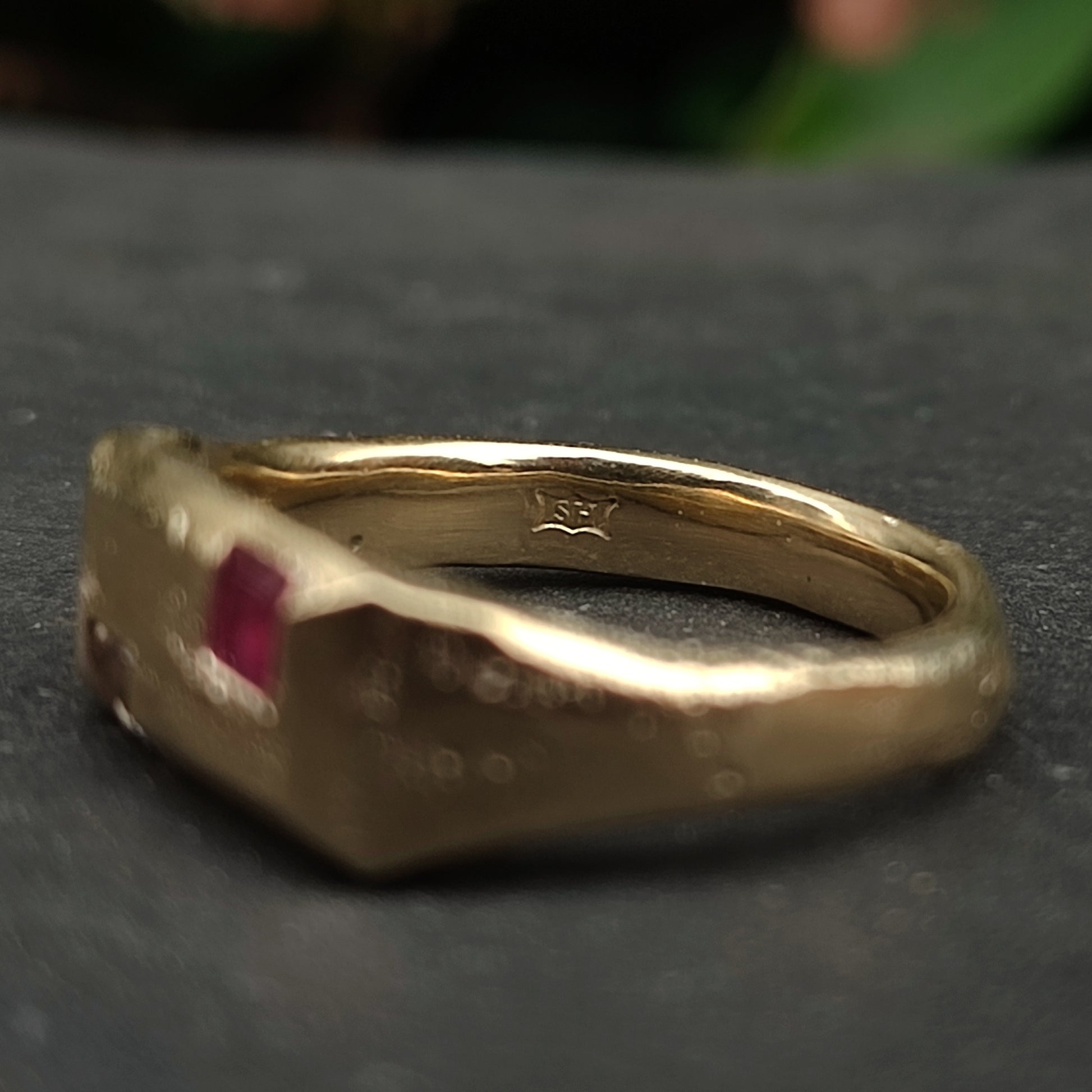 The 9ct Sandcast Sapphire/Ruby Signet by silverhollowjewellery features recycled gold, a rectangular red gemstone, and an engraved "SF" hallmark on the inner band, displayed on a dark surface.