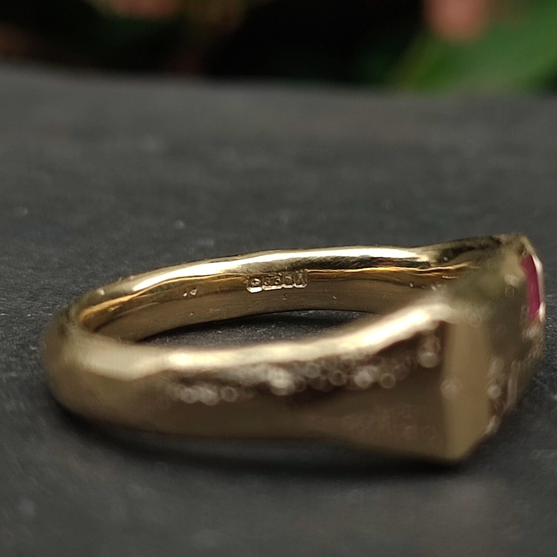 A close-up of the 9ct Sandcast Sapphire/Ruby Signet by silverhollowjewellery, showcasing its sand-cast gold band with a "585" hallmark, set on a dark surface against a blurred green background.