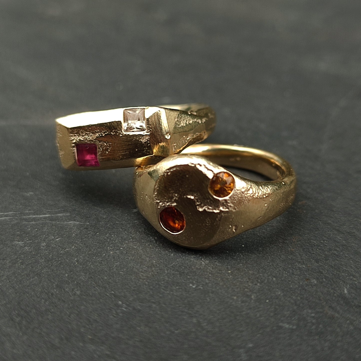 Two gold rings from silverhollowjewellery rest on a dark surface: the 9ct Sandcast Sapphire/Ruby Signet, with square red and clear stones, and another featuring two round orange stones in a yin-yang style.