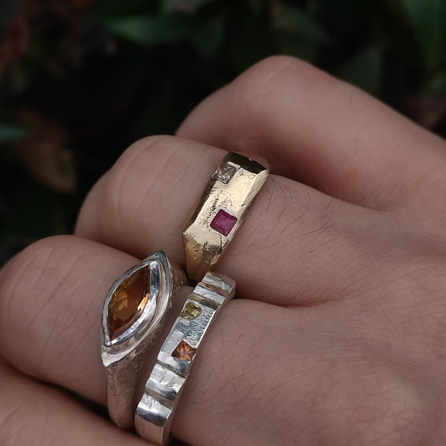 Three fingers display silverhollowjewellery 9ct Sandcast Sapphire/Ruby Signet rings, handcrafted from recycled gold using sand casting, each set with a unique gemstone—amber, yellow, and red—against a blurred green background.