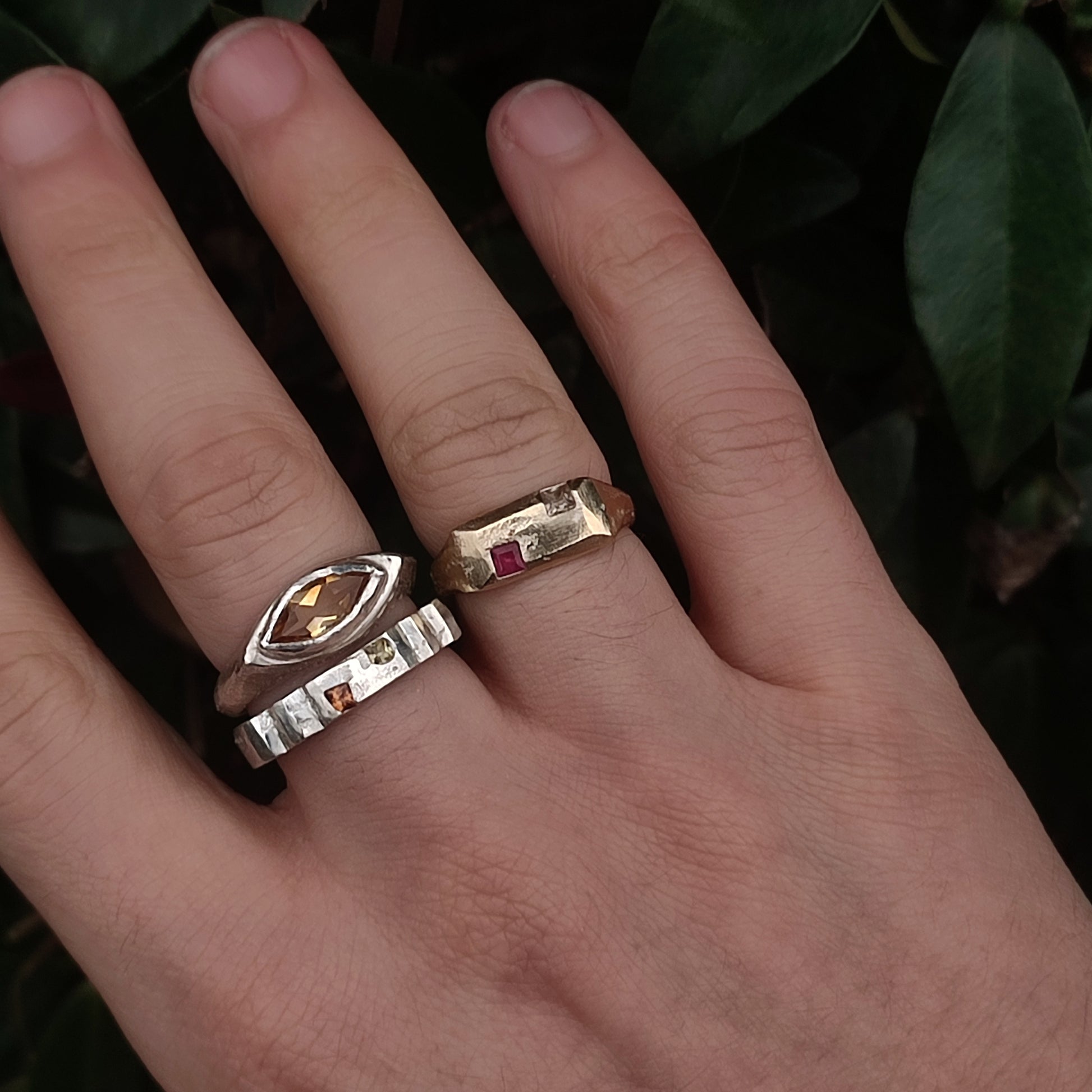 A hand wears three gemstone rings—including silverhollowjewellery’s 9ct Sandcast Sapphire/Ruby Signet in recycled gold with a rectangular red stone—plus two silver rings with white and square stones, set against a background of green leaves.