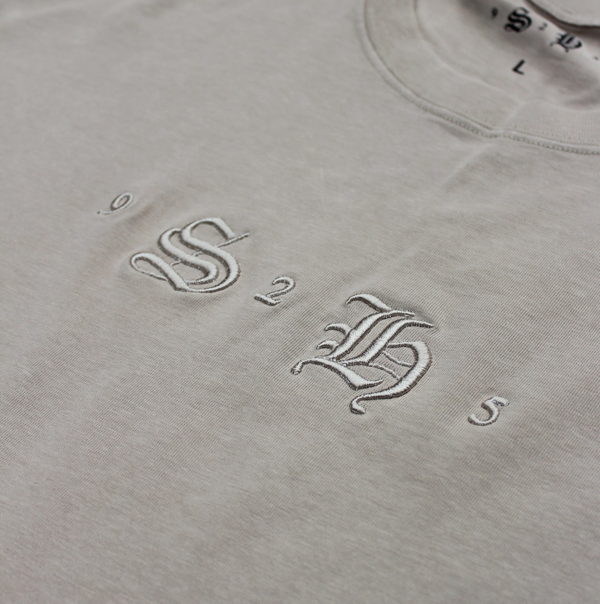 Close-up of the Classic Embroidered T-Shirt by silverhollowjewellery, made from 100% cotton in beige with vintage-style white "9 S B 2 5" embroidery and a visible "L" size label.