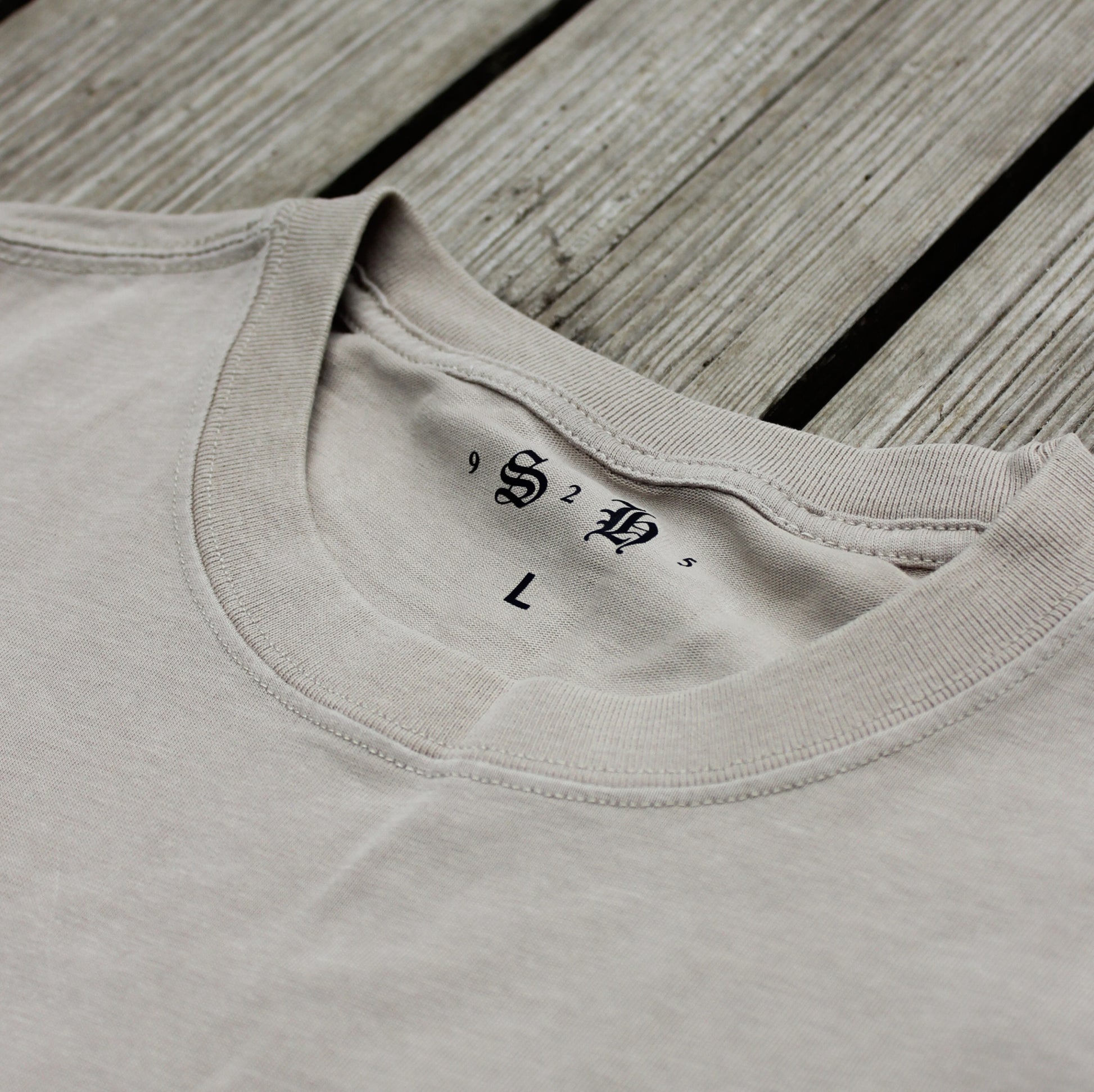 The Classic Embroidered T-Shirt by silverhollowjewellery is a light beige, 100% cotton crew neck in size L, featuring decorative initials inside the collar and shown laid on wooden boards.