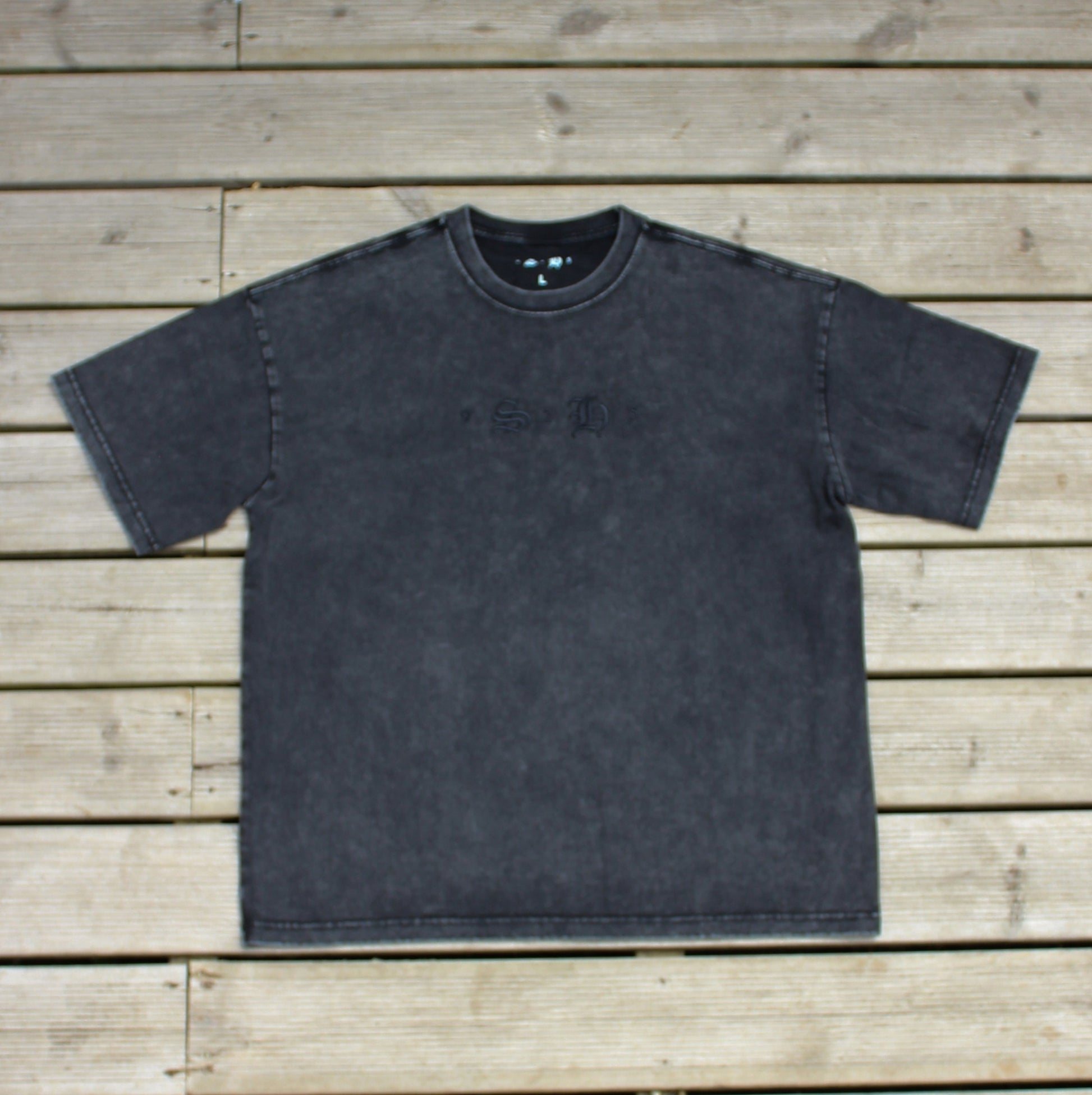 The Classic Embroidered T-Shirt by silverhollowjewellery, a vintage faded black tee with subtle detailing and an embroidered emblem, is displayed flat on a wooden surface.
