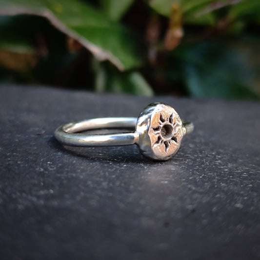The Organic Silver Sun Signet Ring by silverhollowjewellery is a handmade sterling silver piece with a round, textured top featuring a sunburst design, shown resting on a dark surface with blurred green leaves in the background.