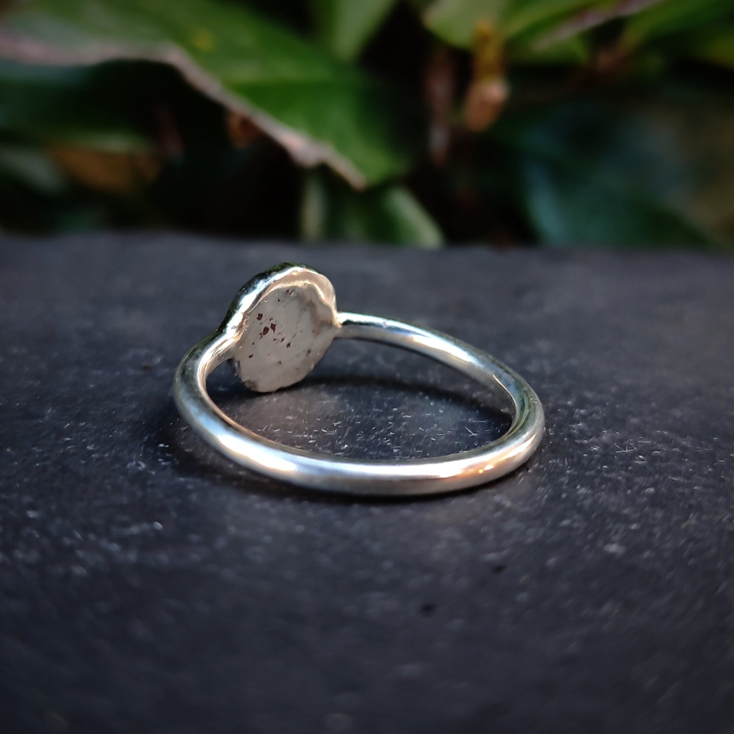 The Organic Silver Sun Signet Ring by silverhollowjewellery, a handmade sterling silver piece with a textured, circular bezel, rests on a dark stone surface with green leaves softly blurred in the background.