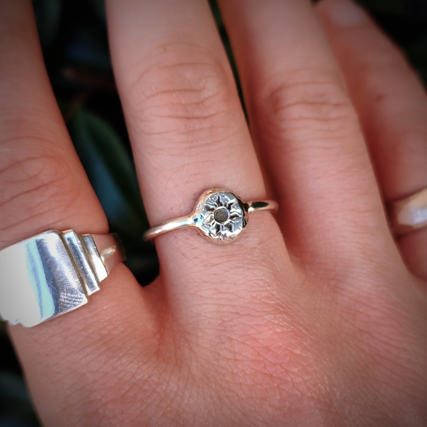 A close-up of a hand wearing the silverhollowjewellery Organic Silver Sun Signet Ring, featuring a round, textured setting with a central dark stone, next to another custom engraved silver band on an adjacent finger.