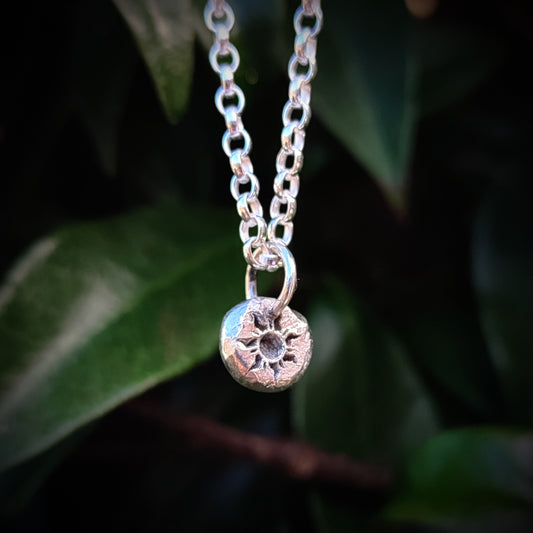 The Organic Silver Sun Pendant by silverhollowjewellery, a sterling silver necklace with a round, textured sun charm, hangs gracefully in front of green leaves.