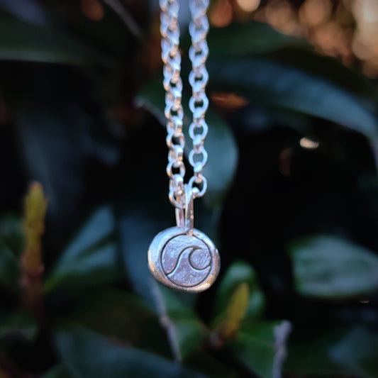 The Organic Silver Wave Pendant by silverhollowjewellery is a sterling silver necklace with a recycled round wave pendant, shown against a blurred green leaf background.