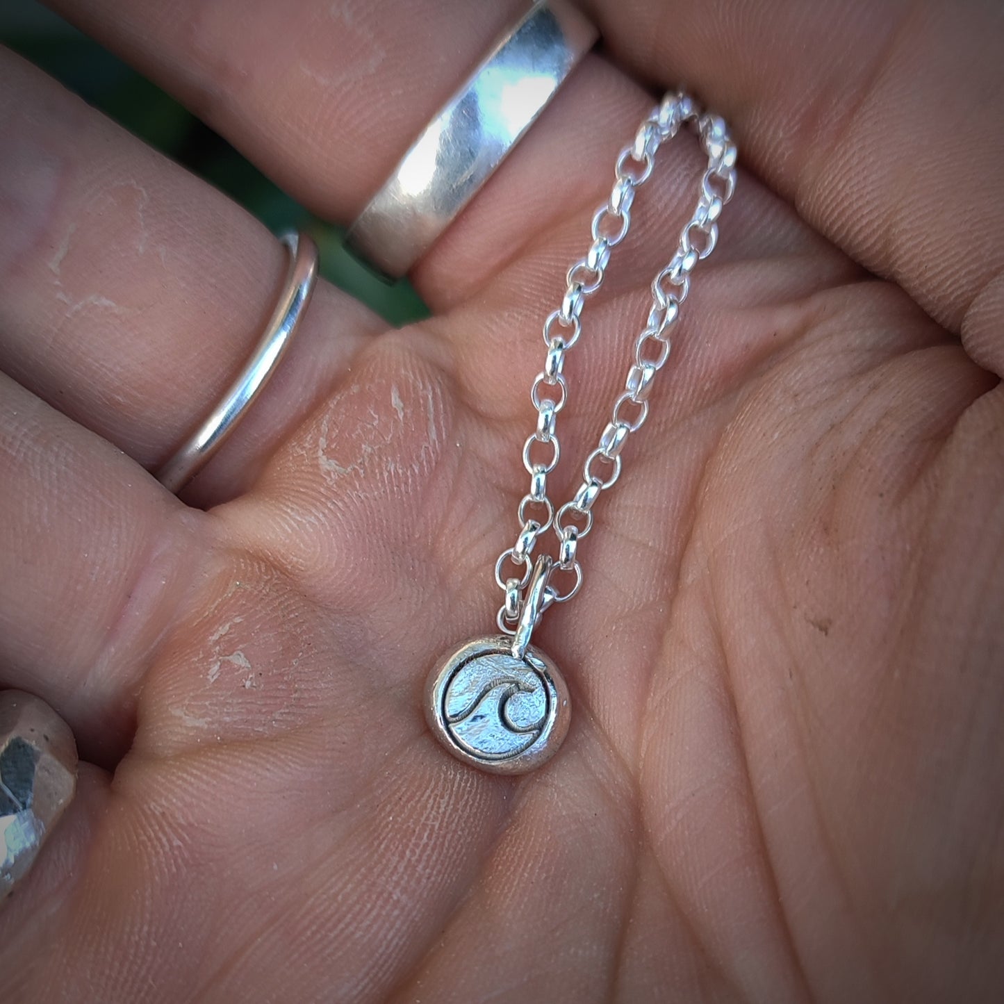 A hand holds the silverhollowjewellery Organic Silver Wave Pendant, a recycled sterling silver chain necklace featuring a small round pendant engraved with a wave design.