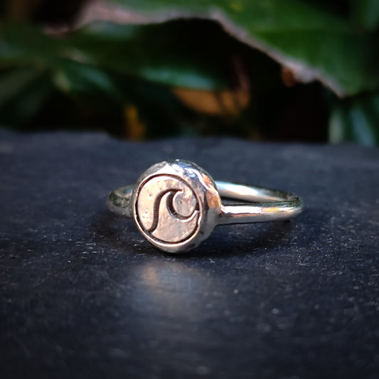 The Organic Silver Wave Signet Ring by silverhollowjewellery, crafted from recycled sterling silver with a round, wave-engraved face, rests on a dark surface accented by green leaves in the background.