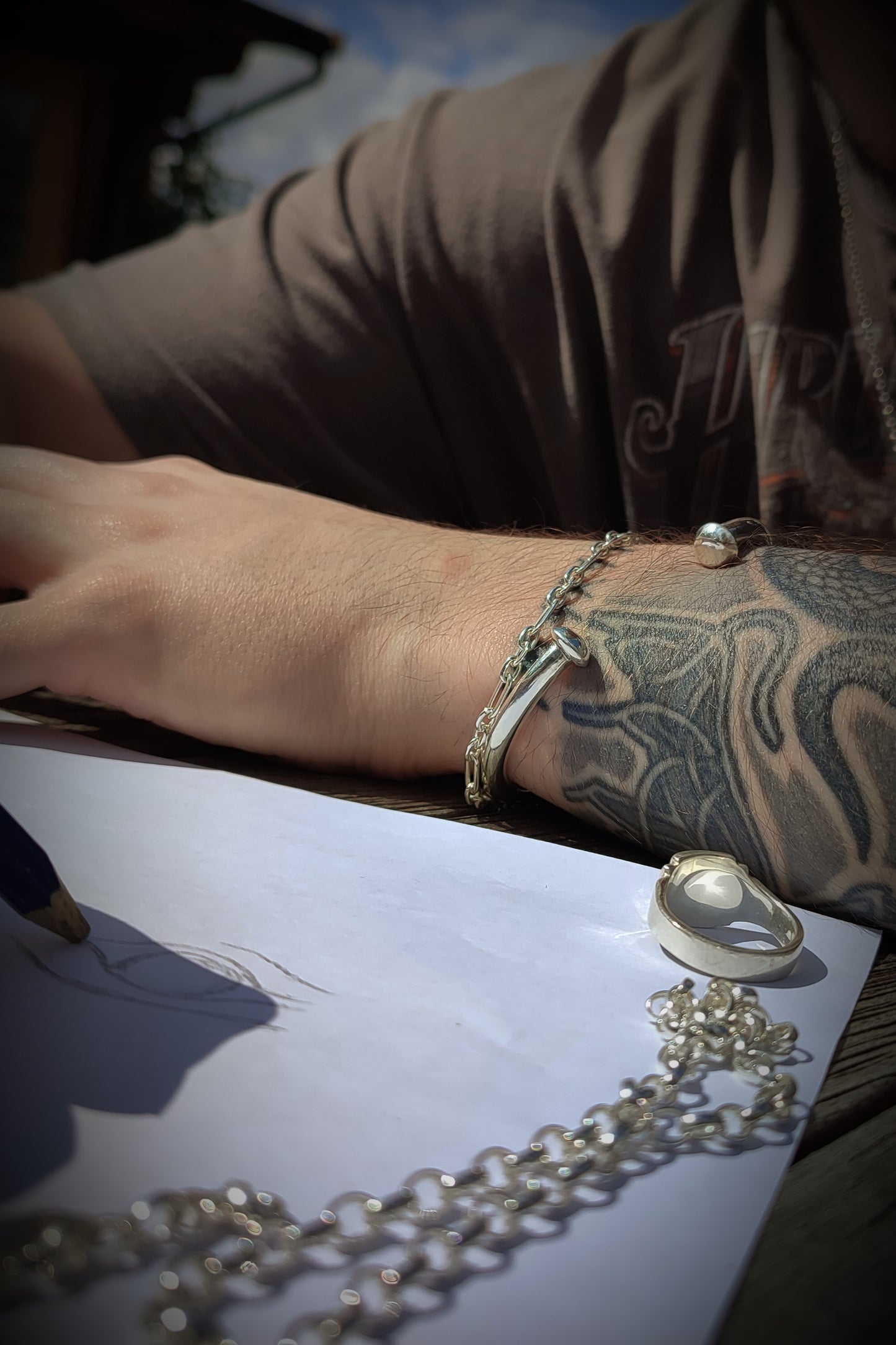A tattooed person wearing the silverhollowjewellery Heavy Silver Bangle sketches on white paper beside a silver necklace and ring on a wooden table.
