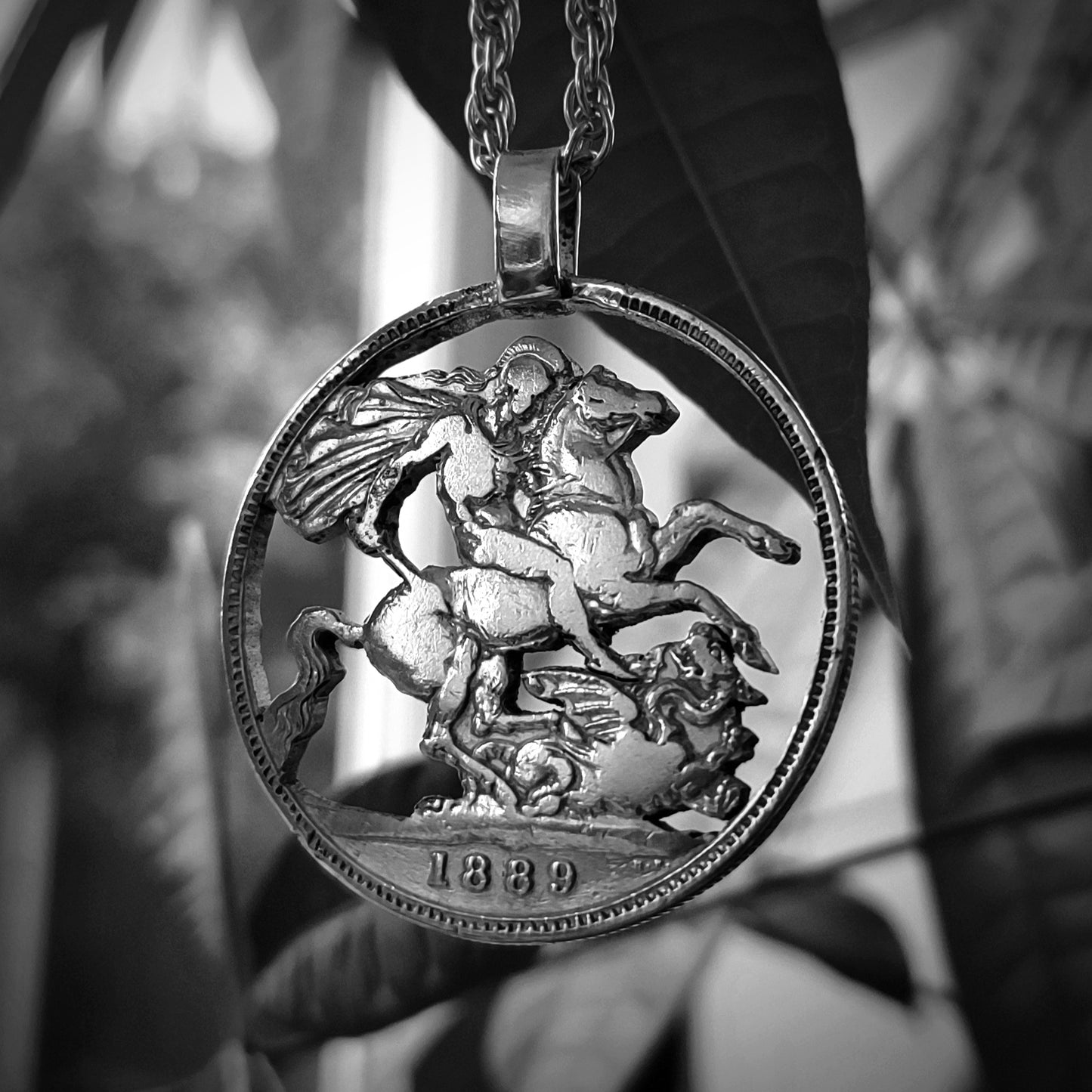 The Sterling Silver Crown Coin Pendant 1800s by silverhollowjewellery hangs in front of leaves, showcasing a detailed relief of a horse and rider slaying a dragon, with "1889" engraved at the bottom.