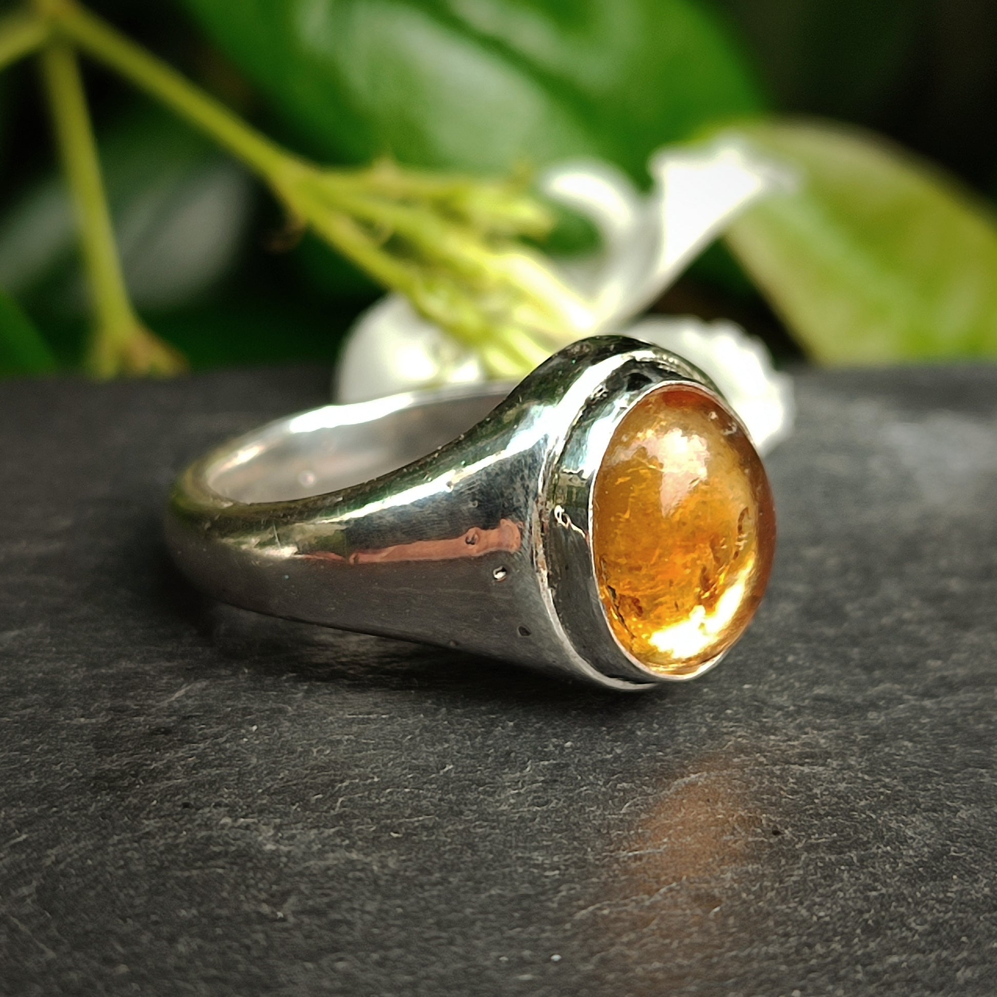 The Citrine Sterling Silver Ring by silverhollowjewellery is displayed on a dark surface, with green leaves and a white flower softly blurred in the background.