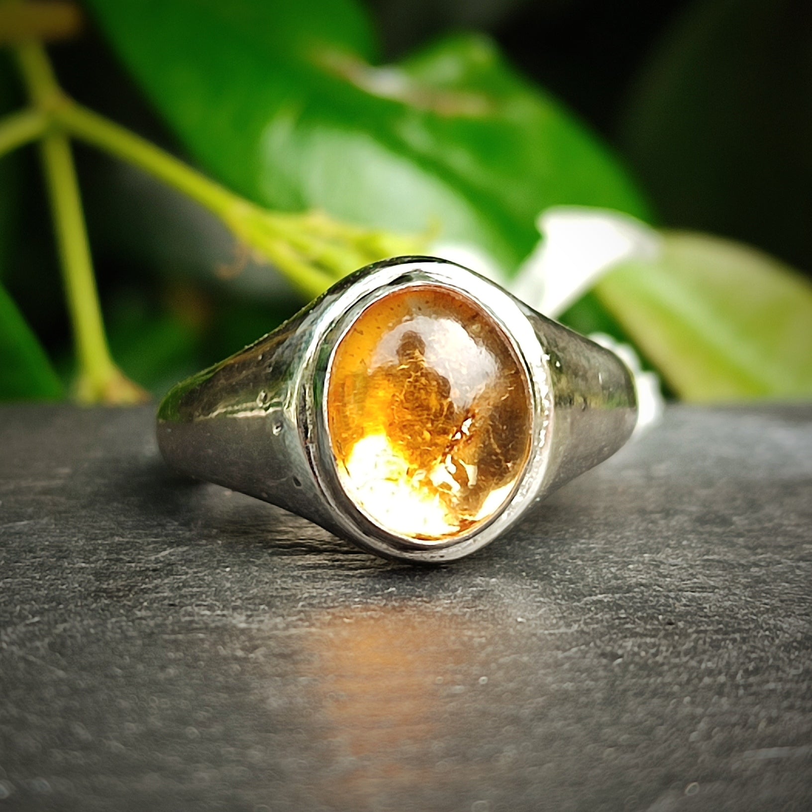 The Citrine Sterling Silver Ring by silverhollowjewellery features a polished citrine gemstone in a sleek sterling silver setting, showcased on a dark surface with green leaves—a gorgeous piece of handcrafted jewelry.