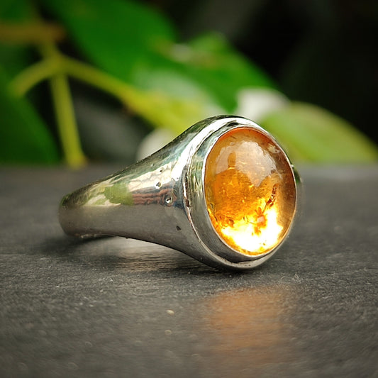 The Citrine Sterling Silver Ring by silverhollowjewellery features a smooth, round citrine gemstone set in the center—an elegant handcrafted piece displayed against a dark surface with green leaves in the background.