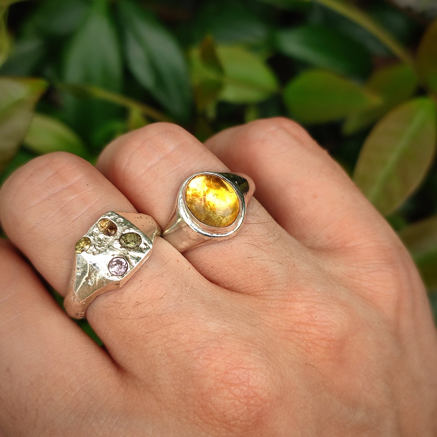 A hand wears two silverhollowjewellery rings—a Citrine Sterling Silver Ring and another with multiple stones—shown against a leafy background.