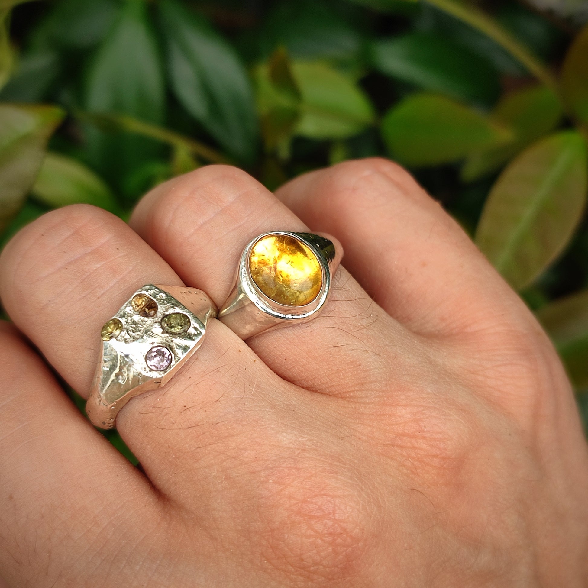 A hand wears two silverhollowjewellery rings—a Citrine Sterling Silver Ring and another with multiple stones—shown against a leafy background.