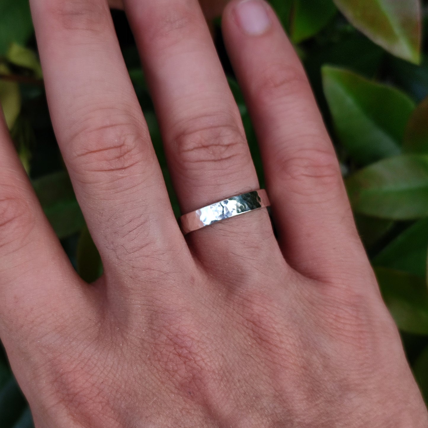 A hand wears the silverhollowjewellery Hammered Ring Thin Sterling Silver, a simple recycled 925 sterling silver band, on the middle finger with green leaves in the background.