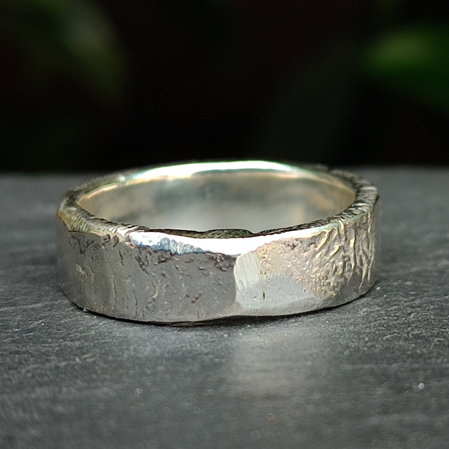 The Custom Distressed Hallmark Ring by silverhollowjewellery features a textured sterling silver design, displayed on a dark surface with a blurred green background—showcasing elegant, eco-friendly jewellery.
