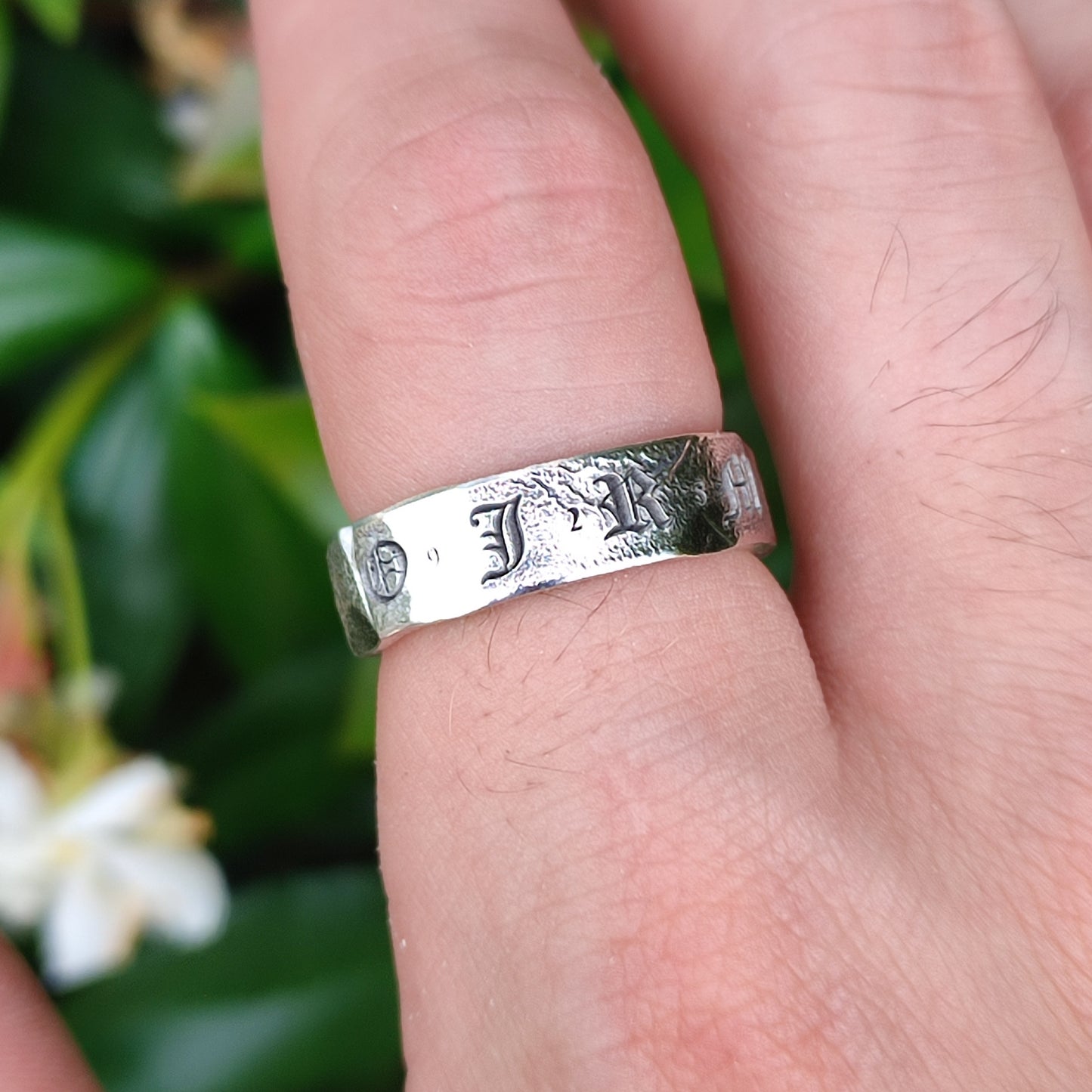 A close-up of a hand wearing silverhollowjewellery's Custom Distressed Hallmark Ring, crafted from recycled sterling silver, with green leaves and white flowers softly blurred in the background.
