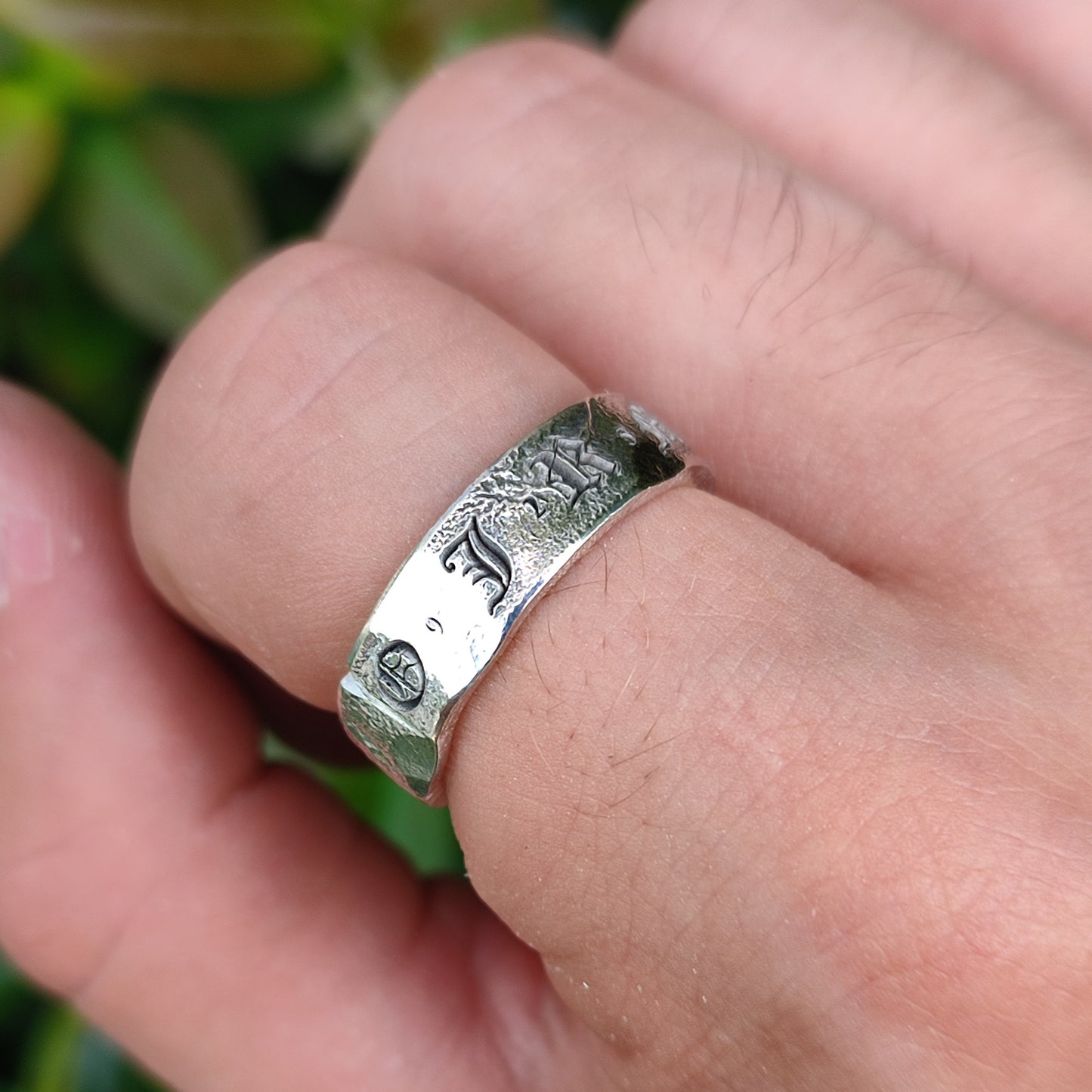 A hand displays the Custom Distressed Hallmark Ring by silverhollowjewellery—an engraved sterling silver piece with an animal motif—highlighted against a blurred green backdrop to showcase eco-friendly style.