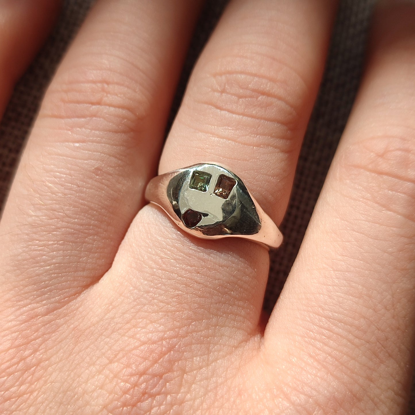 A close-up shows a hand wearing silverhollowjewellery's Sapphire, Garnet and Tourmaline Signet - One Off, a handcrafted sterling silver ring featuring three small square-cut colored stones set at the center.
