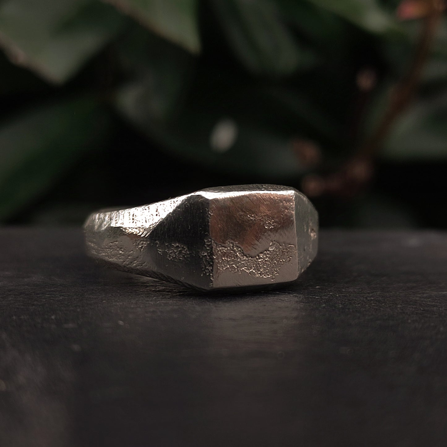 A close-up of the Raw Square Signet by silverhollowjewellery, an organic textured sterling silver ring, resting on a dark surface with blurred green leaves in the background.