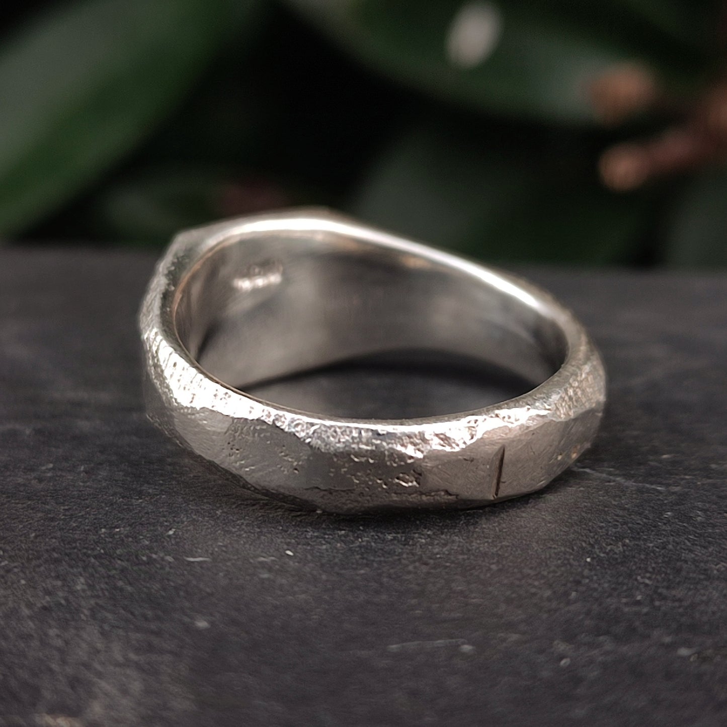 A close-up of the silverhollowjewellery Raw Square Signet, an organic textured sterling silver ring with a rustic uneven surface, resting on a dark surface with green foliage in the background.