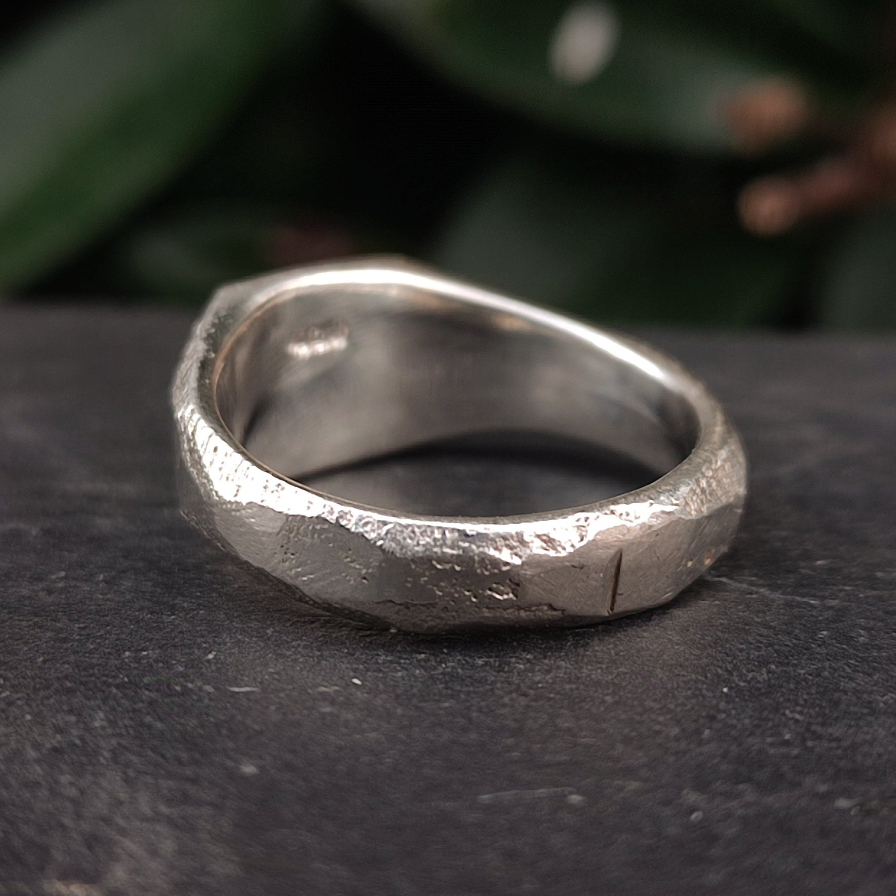 A close-up of the silverhollowjewellery Raw Square Signet, an organic textured sterling silver ring with a rustic uneven surface, resting on a dark surface with green foliage in the background.