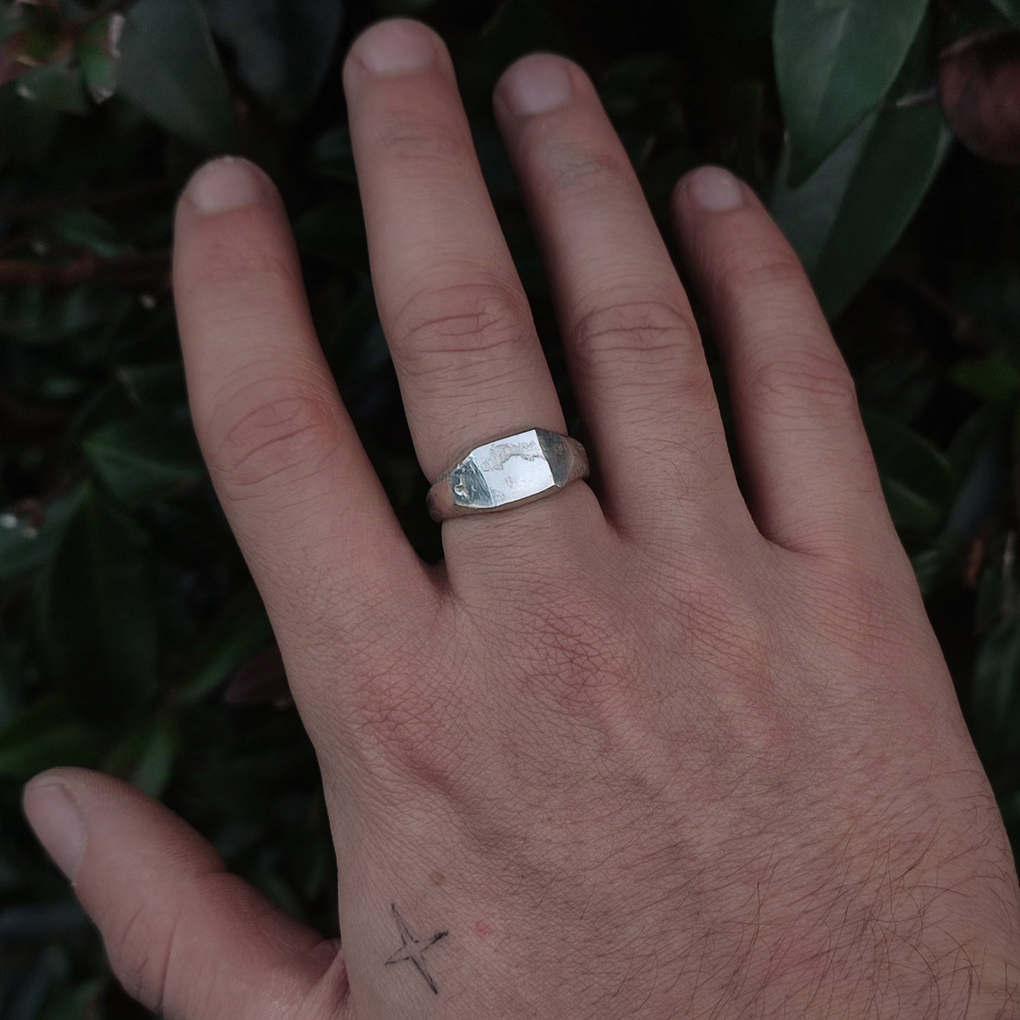 A hand wears the silverhollowjewellery Raw Square Signet, an organic-textured sterling silver ring on the middle finger, with a small cross tattoo by the thumb, set against lush green leaves.