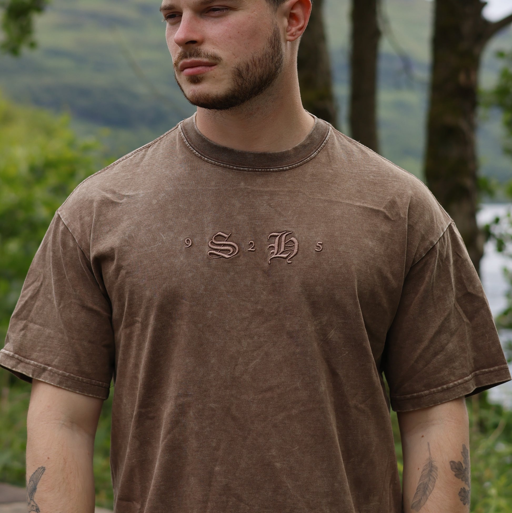 A man in a Classic Embroidered T-Shirt by silverhollowjewellery, featuring "S 2 H" and "9" and "5" on the front, stands outdoors among trees and greenery.