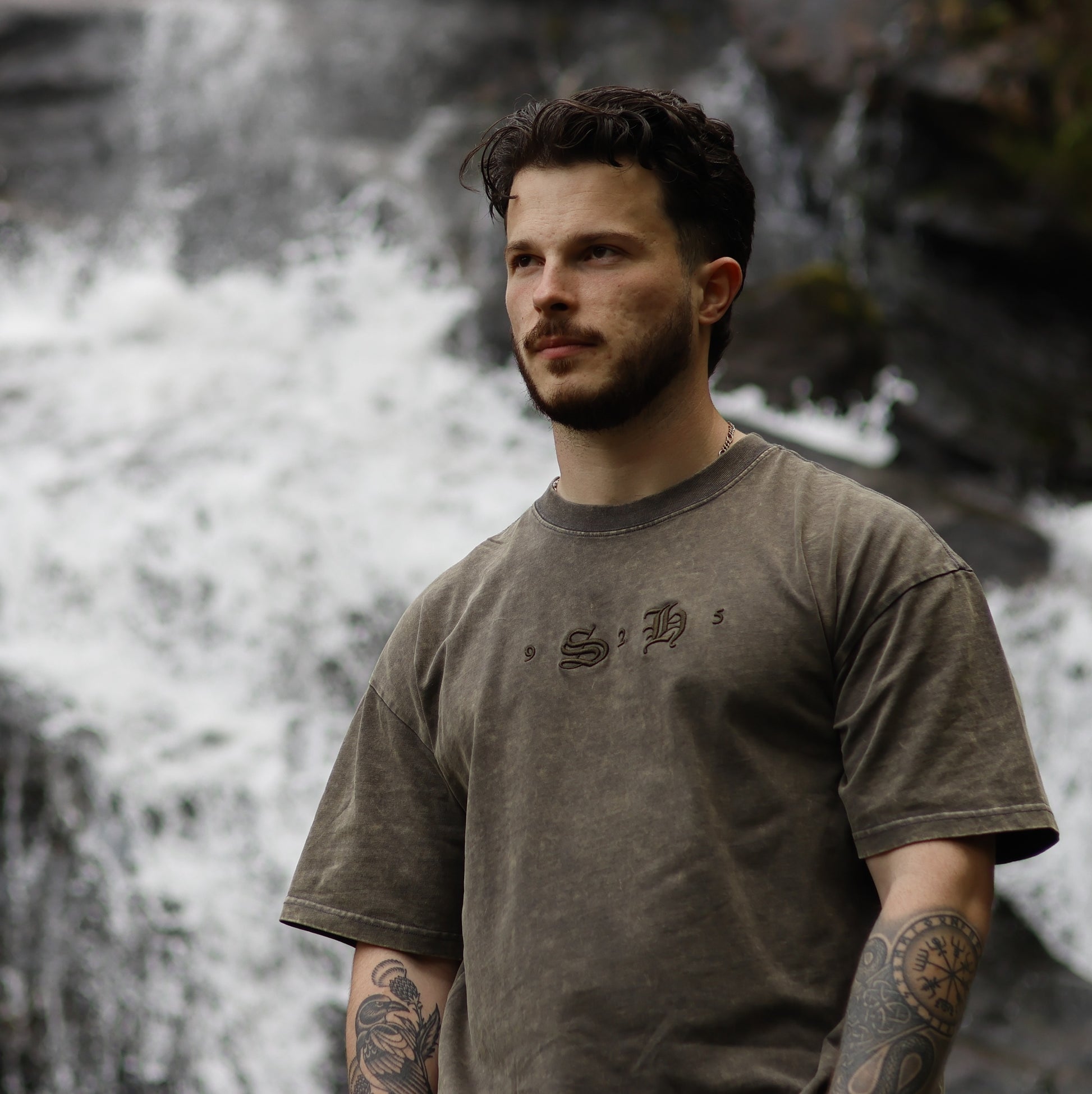 A man with tattooed arms stands before a waterfall, wearing the silverhollowjewellery Classic Embroidered T-Shirt, a faded brown 100% cotton tee with a vintage design, as he gazes off to the side.