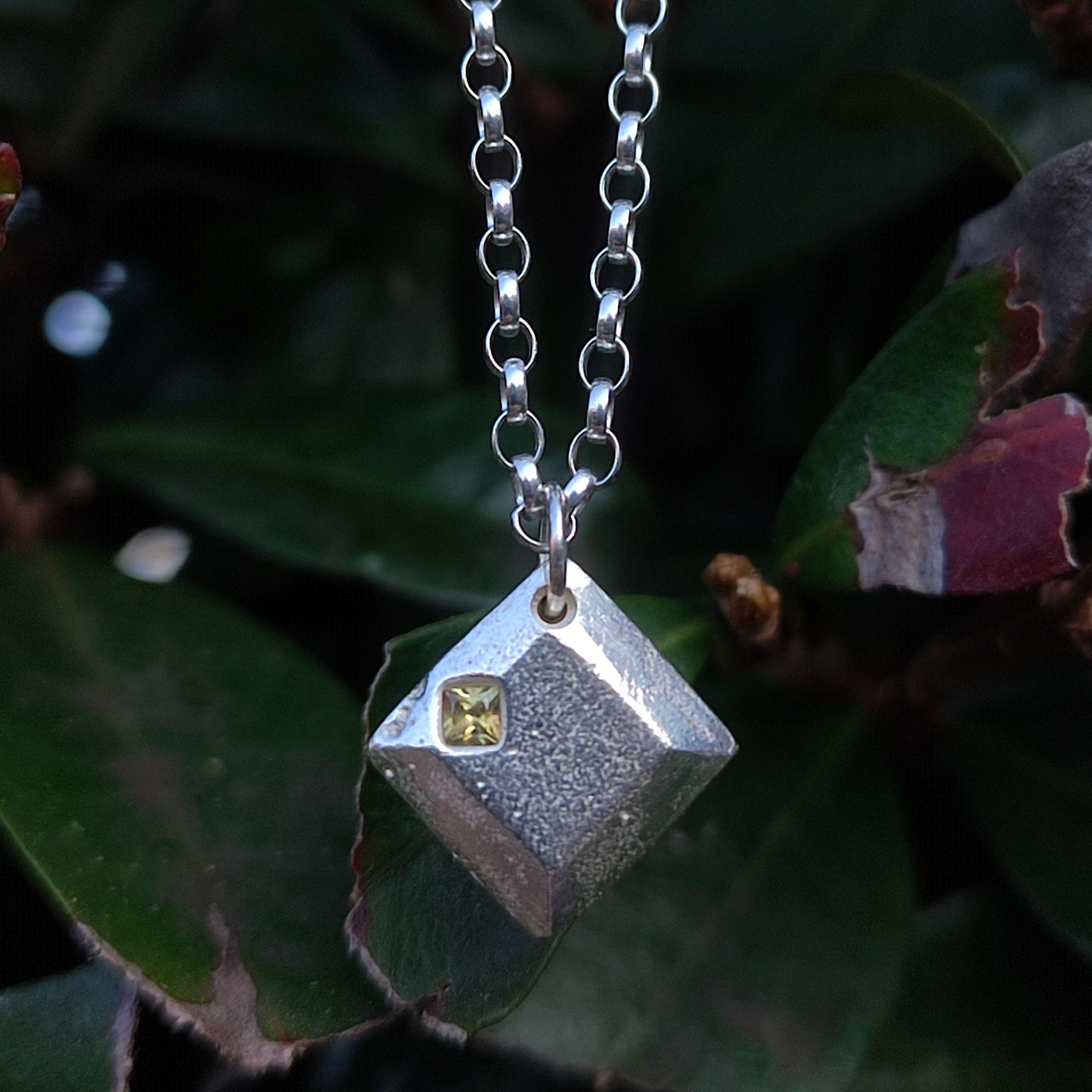 The Sapphire Pendant by silverhollowjewellery features a geometric sterling silver design with a small yellow sapphire, shown hanging from a chain against green leaves.