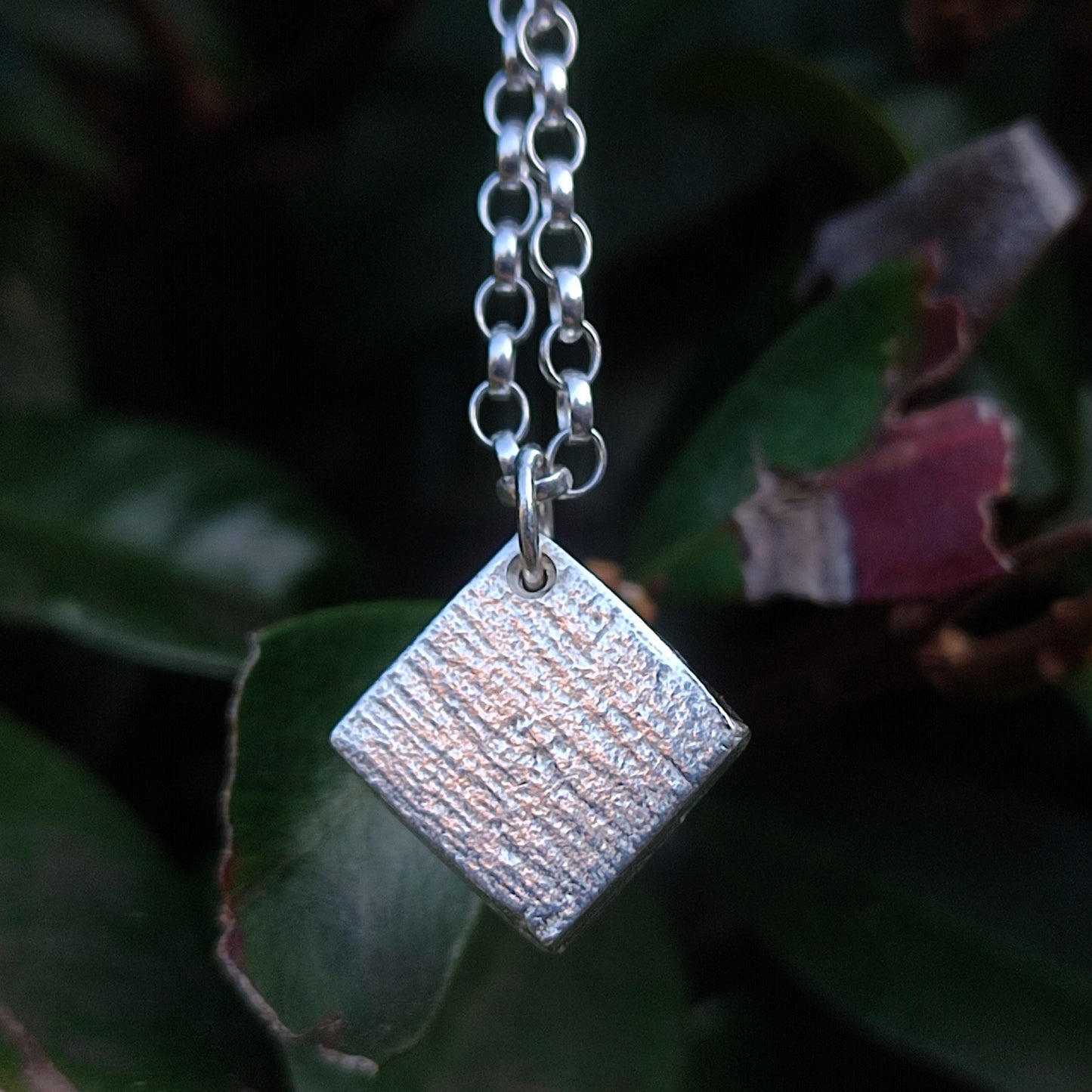 A close-up of the Sapphire Pendant by silverhollowjewellery, crafted from recycled sterling silver with a textured finish, hanging from a chain with green leaves in the background.