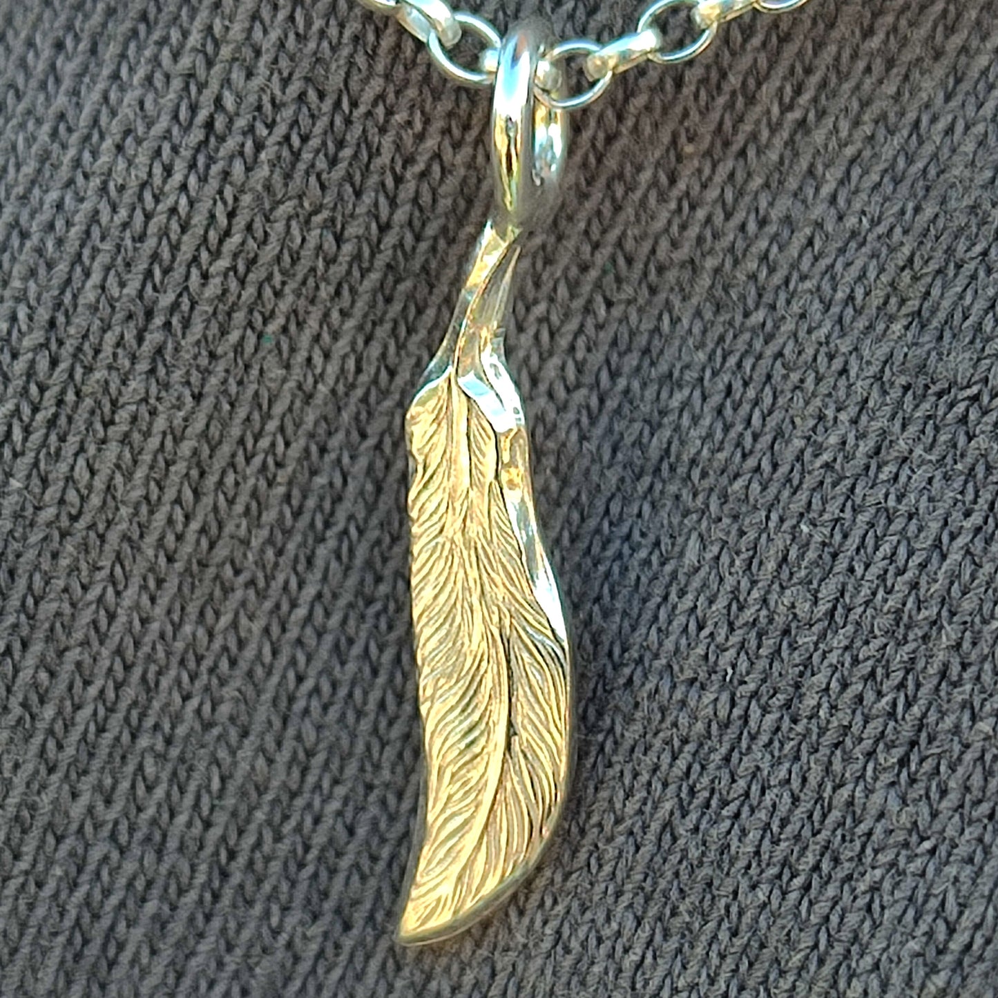 The silverhollowjewellery Feather Pendant-Sterling Silver, crafted from recycled 925 sterling silver in a feather design, hangs on a metal chain against textured gray fabric for a bold unisex necklace look.