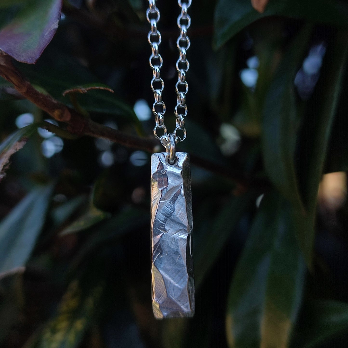 The Rock Pendant by silverhollowjewellery is a rectangular sterling silver piece with a rock-textured surface, crafted from recycled materials and shown hanging from a chain against green leaves.