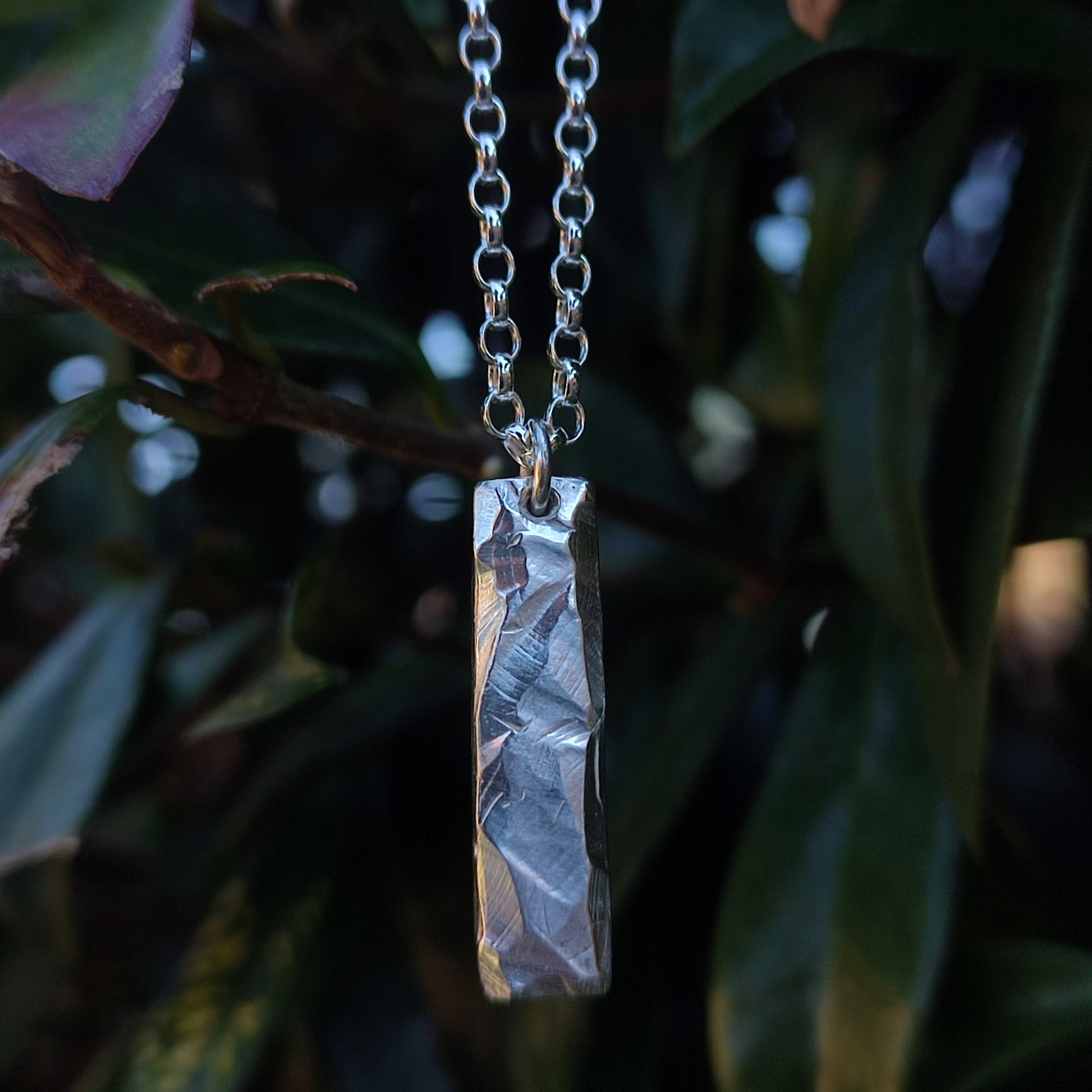 The Rock Pendant by silverhollowjewellery is a rectangular sterling silver piece with a rock-textured surface, crafted from recycled materials and shown hanging from a chain against green leaves.