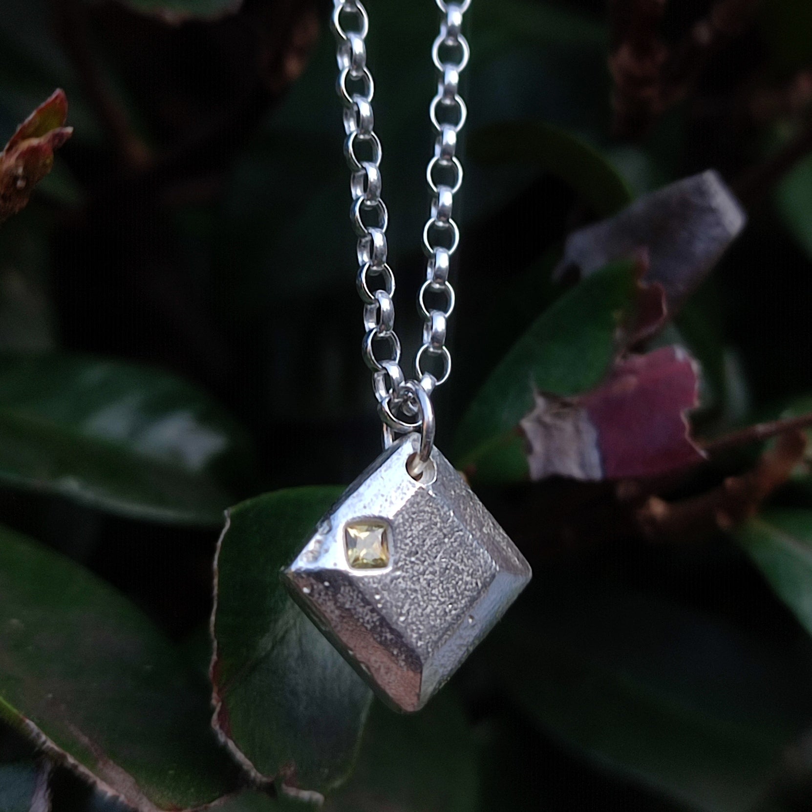 The Sapphire Pendant by silverhollowjewellery features a square, textured recycled sterling silver pendant with a small gold detail on a silver chain, shown hanging in front of green leaves.
