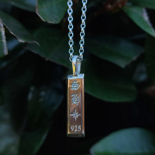 The Makers Mark Pendant by silverhollowjewellery—crafted from recycled sterling silver and engraved with "SH," a star, and "925"—hangs before green leaves, blending eco-friendly design with timeless style.