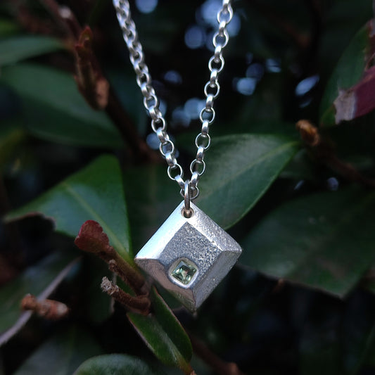 The Sapphire Pendant by silverhollowjewellery hangs on a chain, beautifully photographed against green leaves—an elegant piece of handcrafted jewelry in sterling silver.