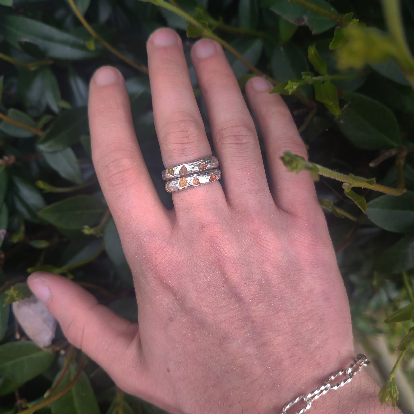 A hand wearing three silverhollowjewellery Raw Sapphire Band Sterling Silver rings, displayed with a backdrop of green leafy plants.