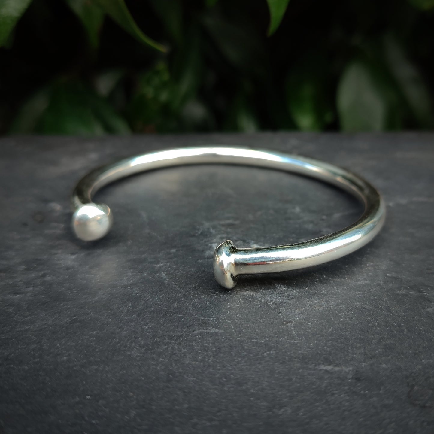 The Heavy Silver Bangle by silverhollowjewellery, crafted from recycled 925 sterling silver with rounded ends, rests on a dark stone surface, accented by green leaves in the background.